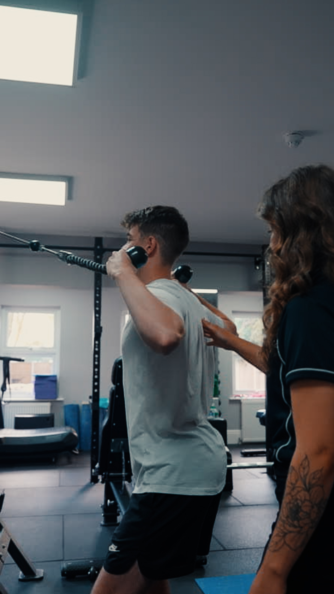 A physiotherapist guiding a client through a cable exercise, hand placed on his back to correct his position.